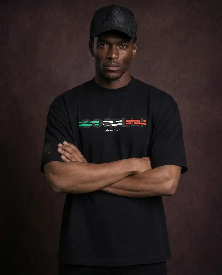 A man wearing a black crewneck t-shirt with a colorful chest graphic and a black baseball cap, standing with arms crossed against a dark brown textured background.