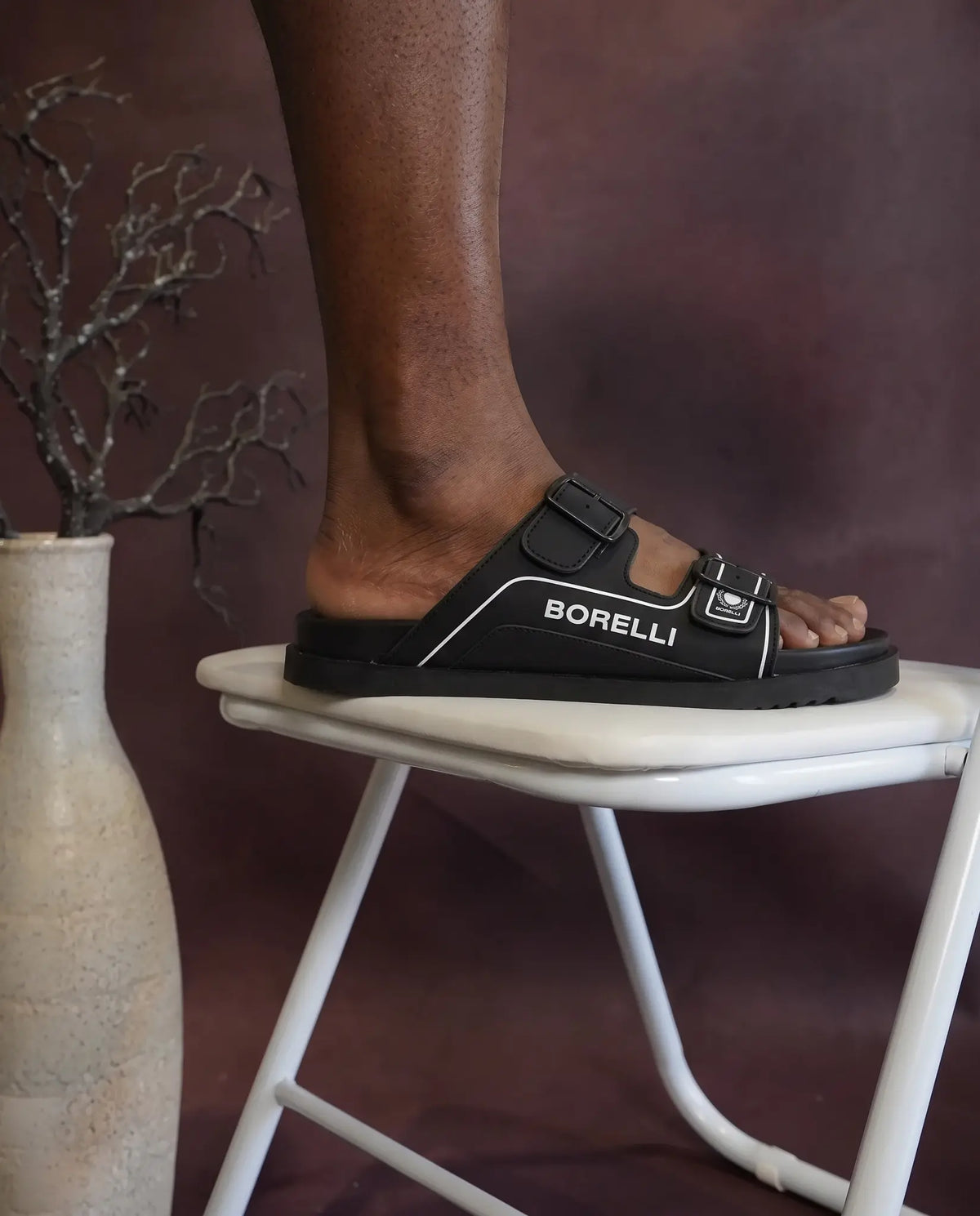 A person wearing a black Borelli double-strap slide sandal while sitting on a white folding stool, with a beige vase and dried branches in the background against a dark brown wall.