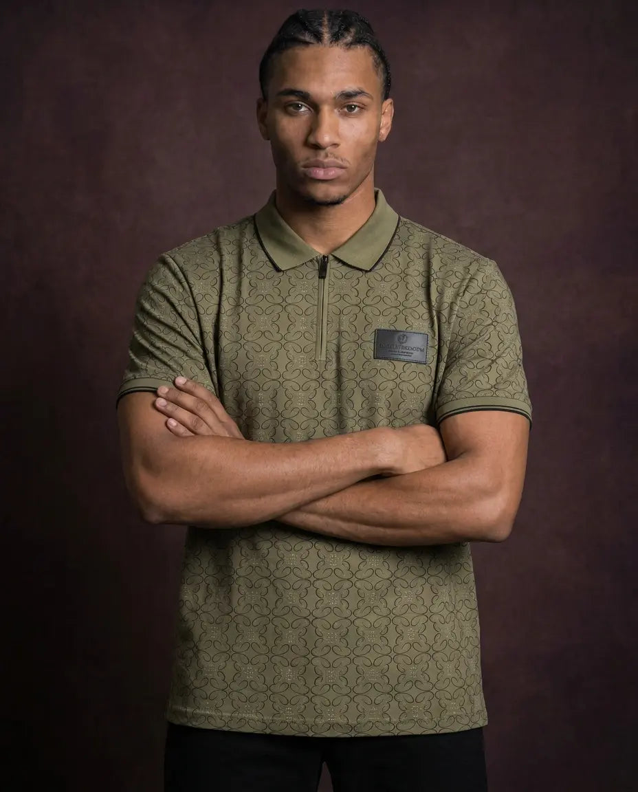 A man wearing a short-sleeve olive green polo shirt with a subtle geometric pattern and a black zipper at the collar, posed with arms crossed against a dark brown studio background.