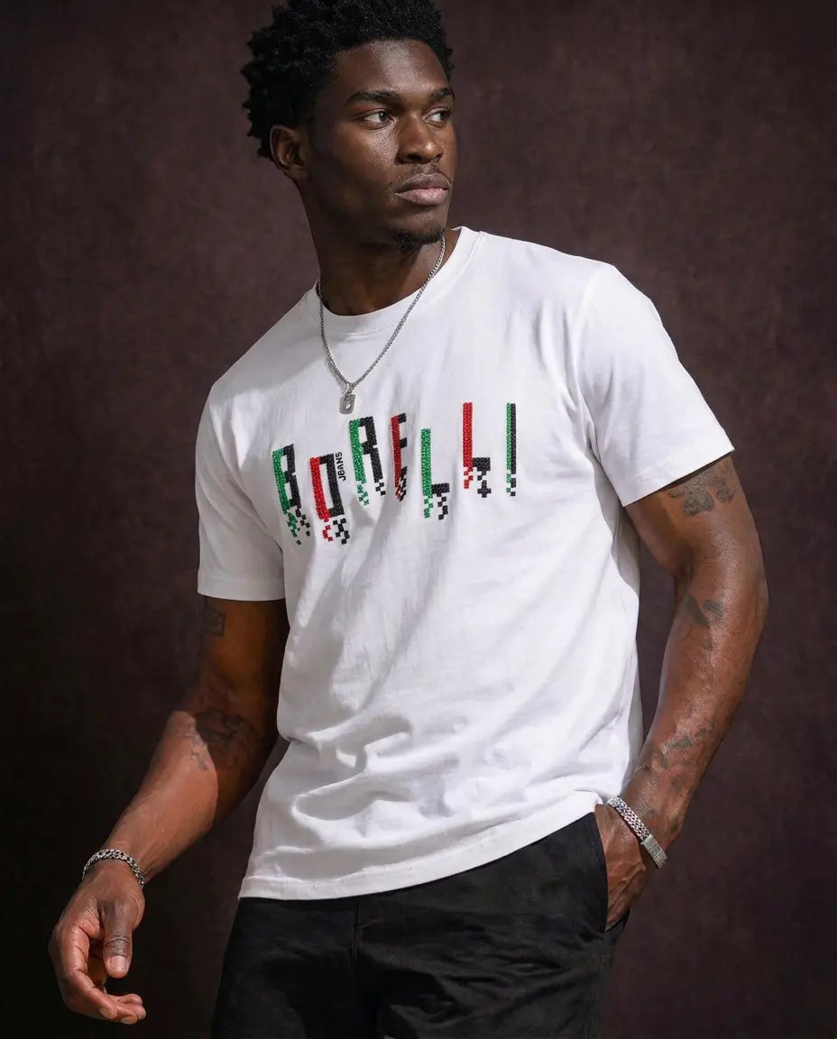 A man wearing a white crewneck t-shirt with 'BORELLI' printed across the chest in a green, red, and black checkered pattern, paired with black pants, set against a dark brown textured background.